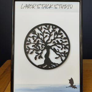 Peut inclure: Œuvre d'art encadrée représentant un arbre noir circulaire avec des branches et des feuilles complexes. L'œuvre est placée sur un fond blanc avec un paysage en bas et le texte "LAYER STACK STUDIO" en haut. Un cadre noir entoure l'œuvre.