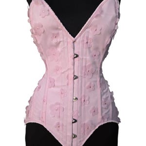 May include: A pink corset with a sweetheart neckline and floral embellishments. The corset has a busk closure in the front and is made of a satin-like fabric.