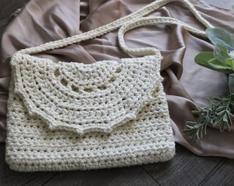 May include: Cream-colored crochet shoulder bag with a flap closure. The bag features a textured stitch pattern and a long, braided strap. The background includes a satin fabric and floral accents, enhancing the handmade aesthetic.