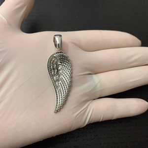 May include: A silver angel wing pendant with a detailed design of small, raised dots. The pendant is on a white surface.