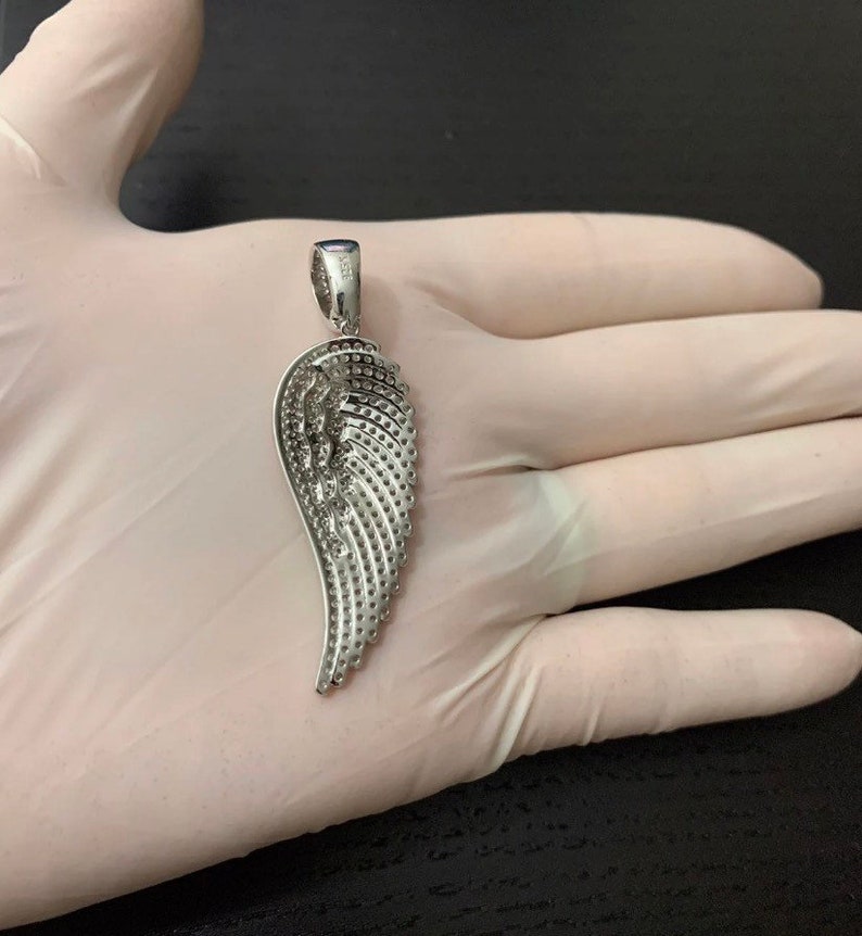 May include: A silver angel wing pendant with a detailed design of small, raised dots. The pendant is on a white surface.