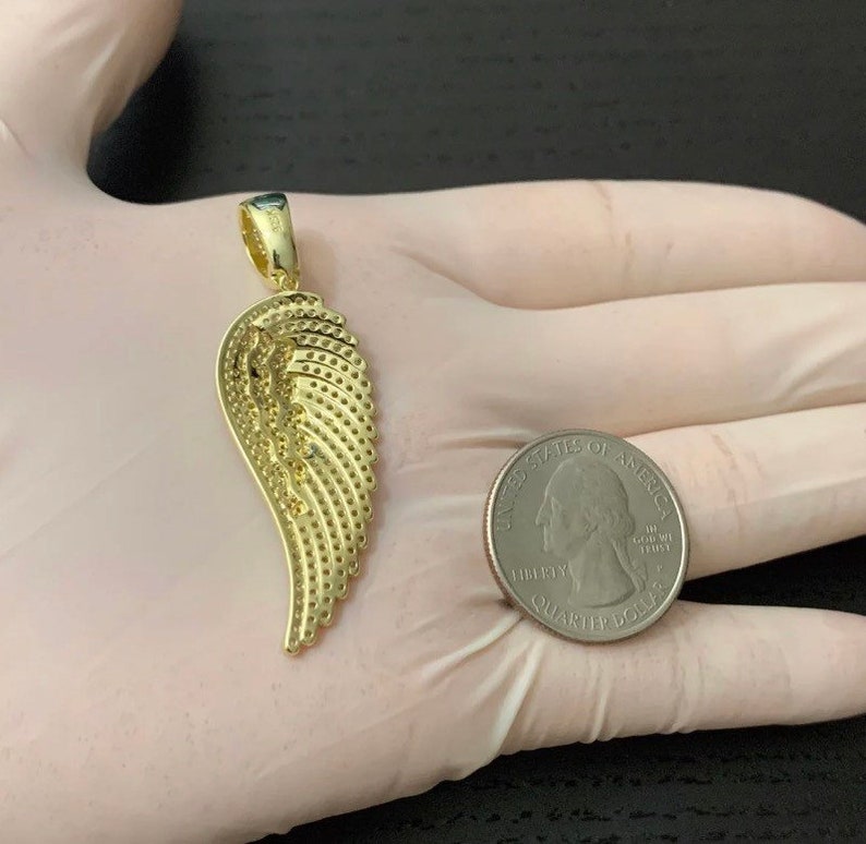 May include: A gold-tone angel wing pendant charm. The wing is detailed with a textured pattern and is shown next to a US quarter for size comparison.