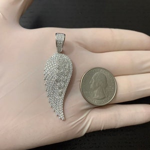 May include: A silver pendant in the shape of a wing, encrusted with small, clear cubic zirconia stones. The pendant is shown next to a US quarter for size comparison.