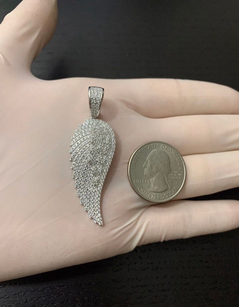 May include: A silver pendant in the shape of a wing, encrusted with small, clear cubic zirconia stones. The pendant is shown next to a US quarter for size comparison.