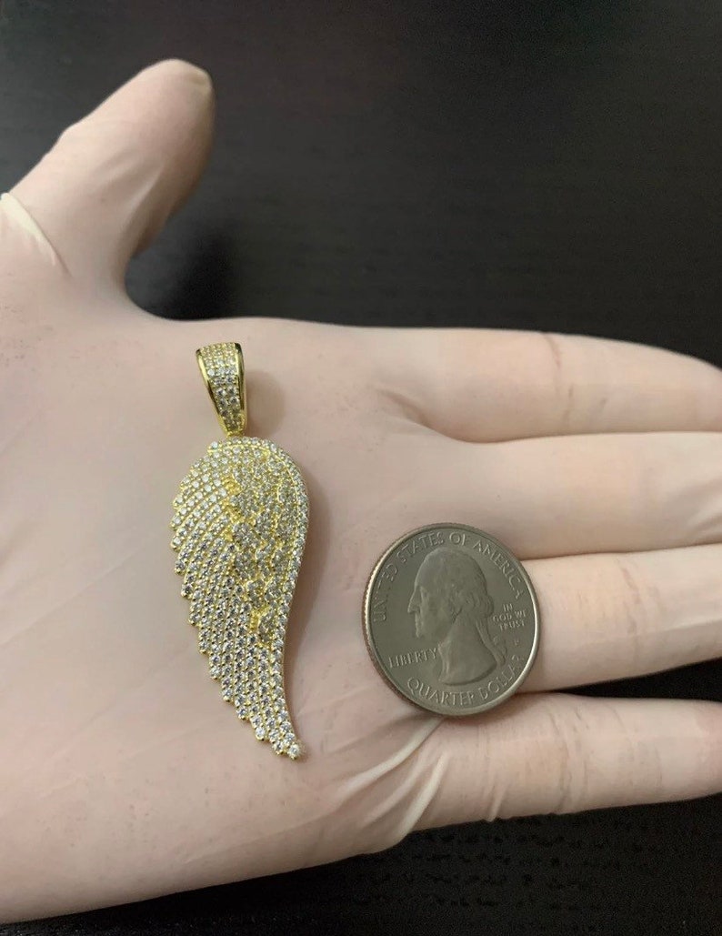 May include: Gold-tone pendant featuring a single angel wing design with clear cubic zirconia stones. The pendant is shown next to a US quarter for size comparison.