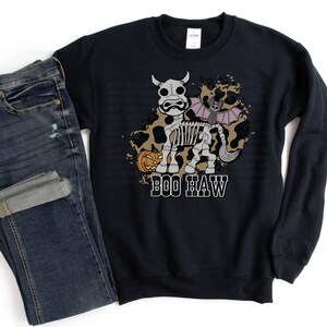 Peut inclure: Sweat-shirt noir avec un motif d'Halloween. Le motif repr&eacute;sente une vache squelette avec une aile de chauve-souris, une citrouille et le texte "BOO HAW" en blanc. Un jean bleu fonc&eacute; est pli&eacute; &agrave; c&ocirc;t&eacute; du sweat-shirt.