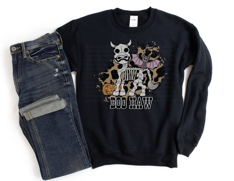 Peut inclure: Sweat-shirt noir avec un motif d'Halloween. Le motif repr&eacute;sente une vache squelette avec une aile de chauve-souris, une citrouille et le texte "BOO HAW" en blanc. Un jean bleu fonc&eacute; est pli&eacute; &agrave; c&ocirc;t&eacute; du sweat-shirt.