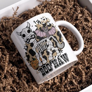 Peut inclure: Mug en c&eacute;ramique blanche avec un motif sur le th&egrave;me d'Halloween. La tasse pr&eacute;sente un squelette de vache de dessin anim&eacute; avec une chauve-souris et une citrouille, et les mots "BOO HAW". La tasse est pr&eacute;sent&eacute;e dans une bo&icirc;te cadeau blanche remplie de papier froiss&eacute; brun.