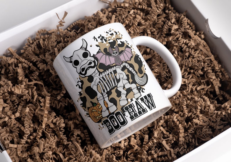 Peut inclure: Mug en c&eacute;ramique blanche avec un motif sur le th&egrave;me d'Halloween. La tasse pr&eacute;sente un squelette de vache de dessin anim&eacute; avec une chauve-souris et une citrouille, et les mots "BOO HAW". La tasse est pr&eacute;sent&eacute;e dans une bo&icirc;te cadeau blanche remplie de papier froiss&eacute; brun.