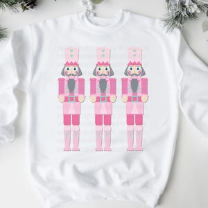 May include: White sweatshirt featuring three pink nutcracker soldier illustrations. Each nutcracker has a pink uniform, a gray beard, and a tall hat. The design is centered on the front of the sweatshirt, perfect for holiday wear.