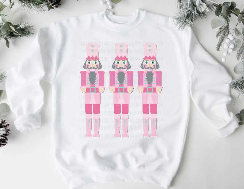 May include: White sweatshirt featuring three pink nutcracker soldier illustrations. Each nutcracker has a pink uniform, a gray beard, and a tall hat. The design is centered on the front of the sweatshirt, perfect for holiday wear.