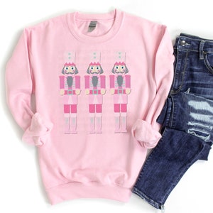 May include: A light pink sweatshirt featuring three cartoon nutcracker figures in pink and gray. The nutcrackers are wearing tall white hats with crowns. Dark blue denim jeans are folded next to the sweatshirt.