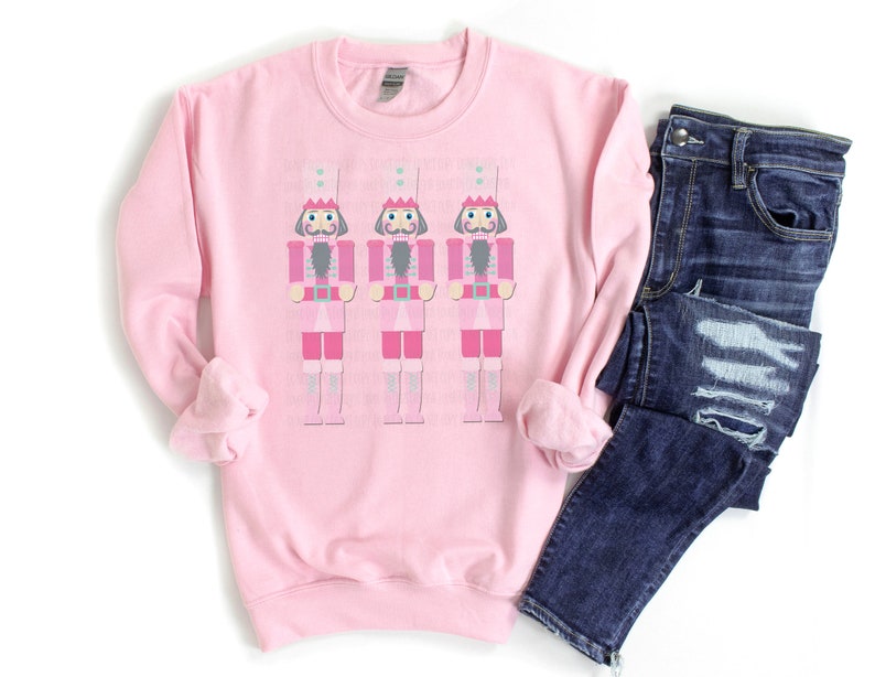 May include: A light pink sweatshirt featuring three cartoon nutcracker figures in pink and gray. The nutcrackers are wearing tall white hats with crowns. Dark blue denim jeans are folded next to the sweatshirt.