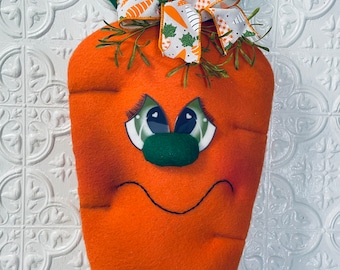 May include: A felt carrot door hanger with a green leafy top, a white and orange bow, and a frowning face with green eyes.