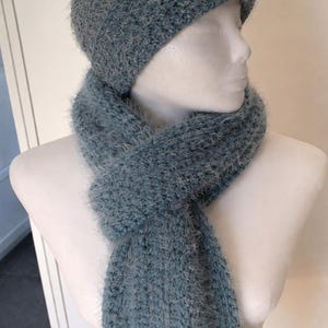 May include: A matching set of a blue-grey knitted hat and scarf. The hat is a close-fitting beanie style. The scarf is long and wrapped around a mannequin's neck. The knitwear has a soft, fuzzy texture.