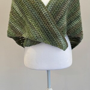 May include: A green and brown crocheted shawl displayed on a white mannequin. The shawl is a wrap style with a V-neck design. The yarn has a variegated color pattern, with shades of green and brown.