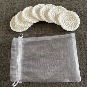 May include: A set of seven white, crocheted, circular facial rounds arranged above a sheer, gray drawstring bag. The rounds are handmade and have a textured appearance, suggesting a reusable skincare product.