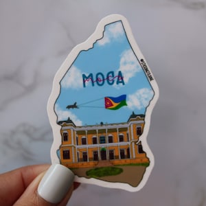 May include: A sticker of a yellow building with a blue sky and clouds. The building has a red roof and a flag flying from the top. The sticker is shaped like the Dominican Republic and says "MOCA" in pink and blue letters.