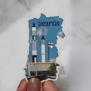 May include: A sticker shaped like the outline of Puerto Rico with a blue sky background. Two rockets are in the foreground, one red and white and the other black and white. The text "BAYAMÓN" is printed in blue at the top of the sticker.