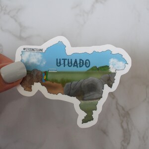 May include: A sticker of the outline of the town of Utuado, Puerto Rico, with a blue sky and clouds. The sticker has the text "Utuado" in blue letters.