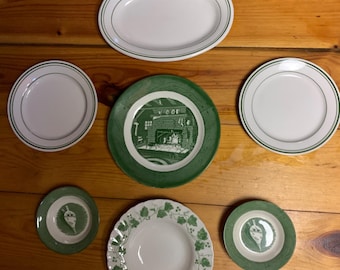 May include: Set of seven green and white ceramic plates with various designs. The plates feature a green rim and a white center with a green design. The designs include a fireplace scene, a vine pattern, and a simple green rim.