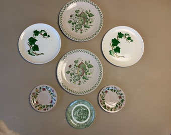May include: A collection of seven white ceramic plates with green and brown floral designs. The plates are arranged in a circle on a beige wall.