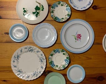 May include: A collection of decorative plates with various floral and leaf patterns. The plates are in shades of blue, green, and white, and some have gold accents. The plates are arranged on a wooden surface.