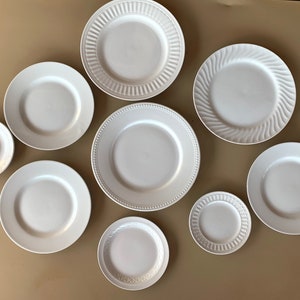May include: A set of nine white ceramic plates of varying sizes and designs. The plates are arranged in a circle on a beige surface.