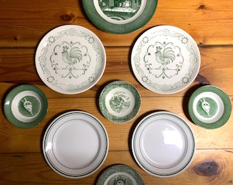 May include: A set of nine green and white ceramic plates with various designs. The plates feature a rooster, a fireplace, a clock, and a house. The plates are arranged on a wooden surface.