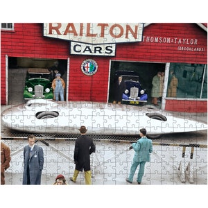 Puede incluir: Rompecabezas completo que presenta una escena vintage en Railton Cars. La imagen muestra un coche de carreras plateado y aerodinámico, coches clásicos en un garaje rojo y personas con ropa de época. El letrero dice "Railton Cars" y "Thomson & Taylor (Brooklands)".