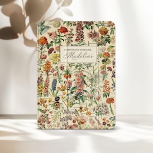 Puede incluir: Un cuaderno de composición con un diseño floral botánico. La portada presenta una variedad de flores y follaje coloridos sobre un fondo crema. El cuaderno tiene el texto "COMPOSITION NOTEBOOK Madeline" en la parte delantera.