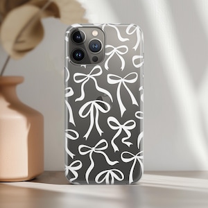 Pode incluir: Capa de telefone transparente com um padrão repetido de laços brancos sobre um fundo cinza escuro. A capa tem um acabamento brilhante e é projetada para um smartphone. Os laços são estilizados e adicionam um toque decorativo ao telefone.