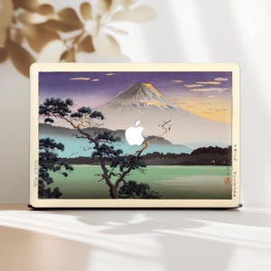Könnte beinhalten: Eine Laptop-Hülle mit einem japanischen Landschaftsdesign, das den Fuji, einen See und einen Kiefernbaum zeigt. Das Kunstwerk verwendet Lila-, Grün- und Gelbtöne mit einem weißen Apple-Logo. Die Hülle hat einen hellbeigen Rand.