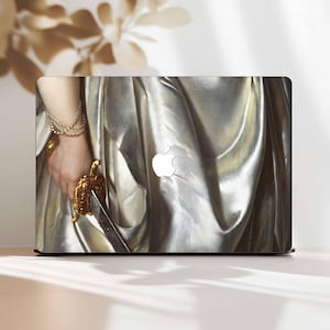 May include: Laptop skin featuring a detail from a classical painting. The design shows a hand holding a golden sword, adorned with a pearl bracelet and ring. The background is a draped, silvery fabric. The Apple logo is visible.