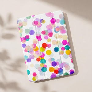 Puede incluir: Una funda rectangular para tableta con fondo blanco cubierta por un patrón disperso de círculos translúcidos y superpuestos en tonos vibrantes de rosa, morado, azul, amarillo, naranja y verde, con un diseño de tres pliegues visible al frente.