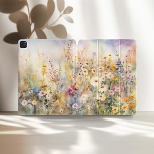 Puede incluir: Una funda para iPad abierta con un diseño floral en acuarela. La funda muestra una vibrante escena de pradera con margaritas, flores silvestres y hierbas en tonos de amarillo, rosa, morado y azul. La funda está diseñada para proteger el iPad.