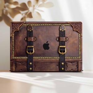 May include: A brown laptop case with a vintage trunk design. The case features decorative gold trim, black straps with gold buckles, and a black Apple logo. The case has a distressed leather appearance.