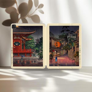 以下が含まれることがあります： オフホワイトのiPadケース。日本の風景画がプリントされており、赤い寺院と雨の降る街並みが描かれ、傘をさす人物がいます。ケースは開いており、内部と詳細なアートワークが示されています。