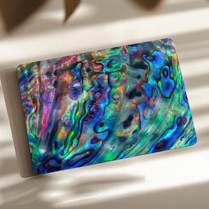 May include: A rectangular abalone shell piece featuring a vibrant, iridescent surface with swirling patterns of deep blue, emerald green, magenta, and gold. The polished material displays a natural, multi-colored luster with organic textures.