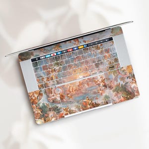 May include: Laptop with a decorative skin featuring a classical painting with cherubs, clouds, and figures in shades of beige, blue, and orange. The keyboard keys are visible, and the laptop is open.