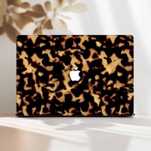 May include: A laptop with a tortoiseshell pattern cover. The cover features a repeating pattern of dark brown and tan shapes. The Apple logo is visible in white on the laptop's surface. The laptop is on a white surface.