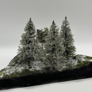 Puede incluir: Una escena de invierno en miniatura con tres pequeños árboles de hoja perenne cubiertos de nieve blanca sobre una base negra. Los árboles están dispuestos en forma triangular con una pequeña roca en el fondo.