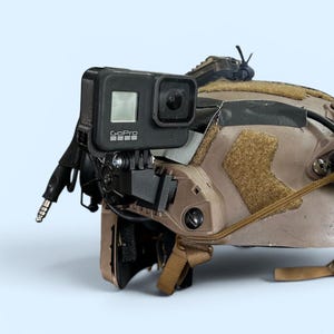 May include: A tan tactical helmet with a mounted black GoPro camera. The helmet has various attachments, straps, and wiring. The camera is positioned on the front of the helmet, with a small screen visible. The background is a light blue.