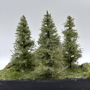 May include: A miniature landscape with three model evergreen trees. The trees are a muted green colour and are set on a small, grassy hill with a black base, creating a diorama scene.