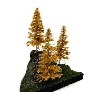 Könnte beinhalten: Miniatur-Diorama mit drei goldgelben Modellkiefern auf einer kleinen, strukturierten, dunkelgrünen und schwarzen Basis. Die Basis hat ein felsiges, moosiges Aussehen und schafft eine kleine Waldszene vor weißem Hintergrund.