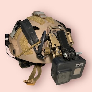 May include: A tan helmet with a black strap and a GoPro camera mounted on the side. The camera is attached to a black and gold mount.