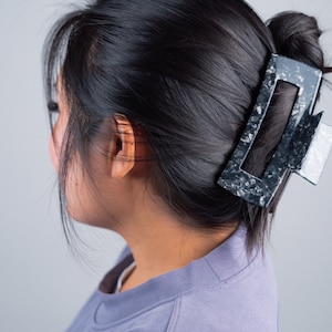 May include: A woman with long black hair is wearing a purple shirt. Her hair is pulled back with a large black and white hair clip.