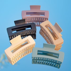 May include: Five rectangular hair claw clips in various pastel colors: light pink, black, cream, beige, and blue. The clips have a matte finish and a curved design with teeth for gripping hair. They are arranged on a reflective surface against a blue background.
