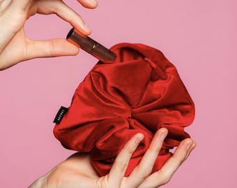 May include: A red velvet scrunchie with the word "SCRUNCH" printed on the side. A hand is holding the scrunchie and placing a gold lipstick tube inside.