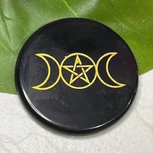 May include: A circular black stone disc featuring a gold-colored engraved triple moon symbol with a central pentagram. The design consists of two crescent moons flanking a circle containing a five-pointed star, all rendered in thin gold lines.